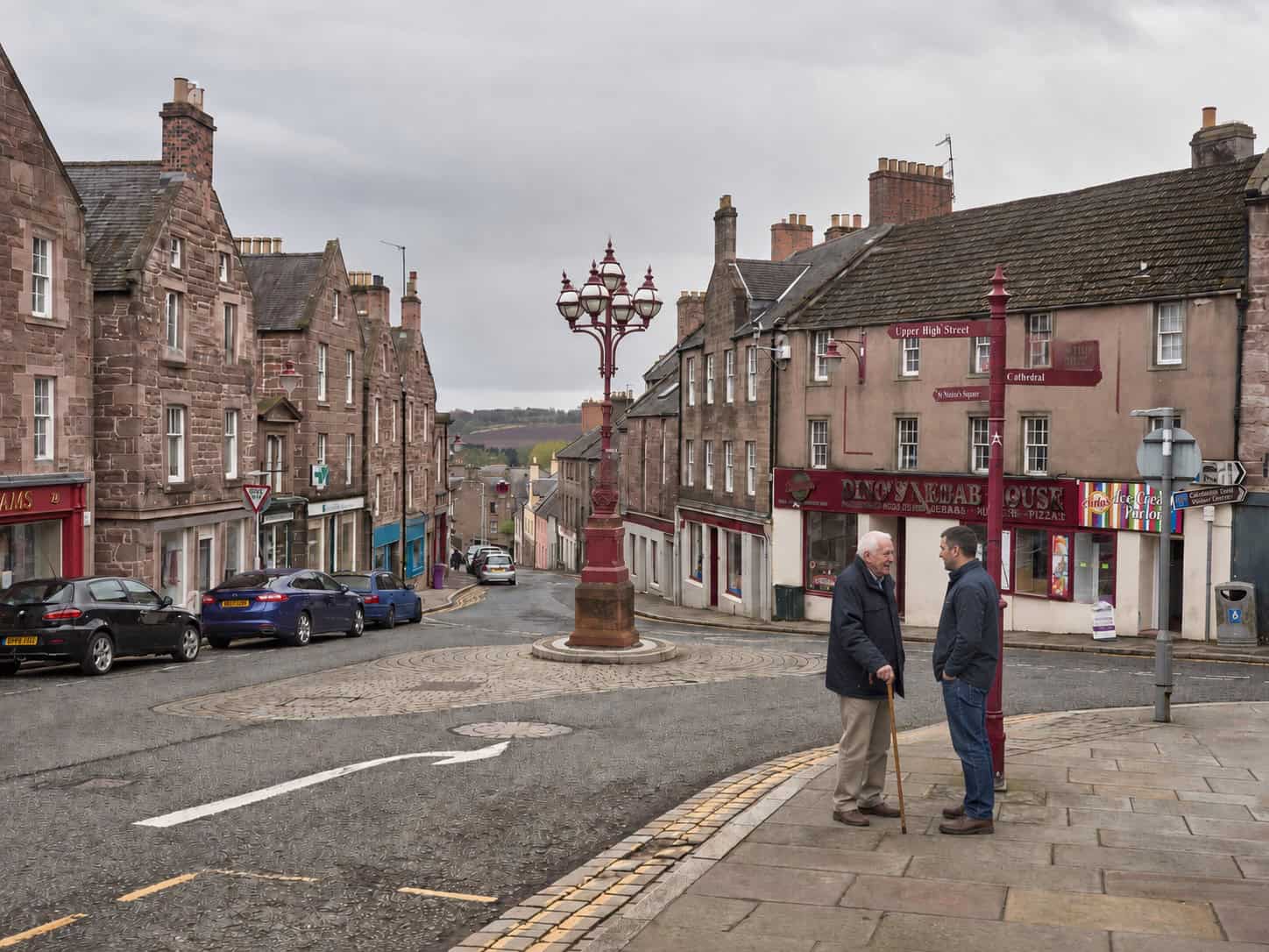 Two men talking in a historic Scottish town square with old buildings and a decorative street lamp.